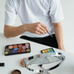 Close-up of a person applying makeup with a colorful eyeshadow palette and brush on a white table.